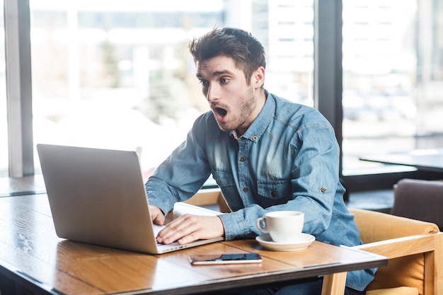 ¡No creas! El retrato de vista lateral de un joven hombre de negocios conmocionado emocionalmente con pantalones de jeans azul está sentado en un café, leyendo noticias y trabajando de forma remota con grandes ojos sorprendidos mirando a la computadora portátil.