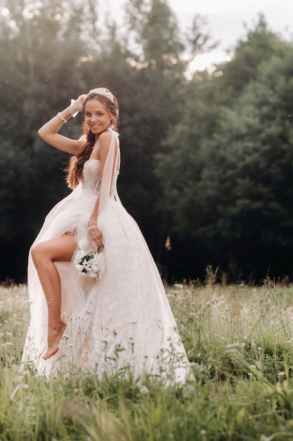 No dia do casamento, uma noiva elegante em um vestido longo branco e luvas com um buquê nas mãos está em uma clareira curtindo a natureza. bielo-rússia.