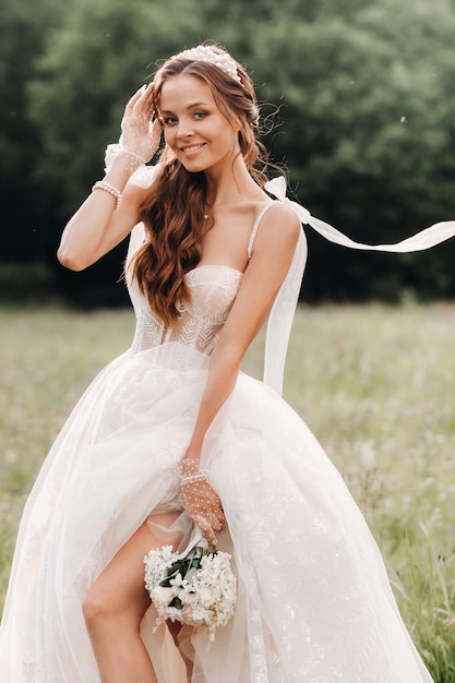 No dia do casamento, uma noiva elegante em um vestido longo branco e luvas com um buquê nas mãos está em uma clareira curtindo a natureza. Bielo-Rússia.