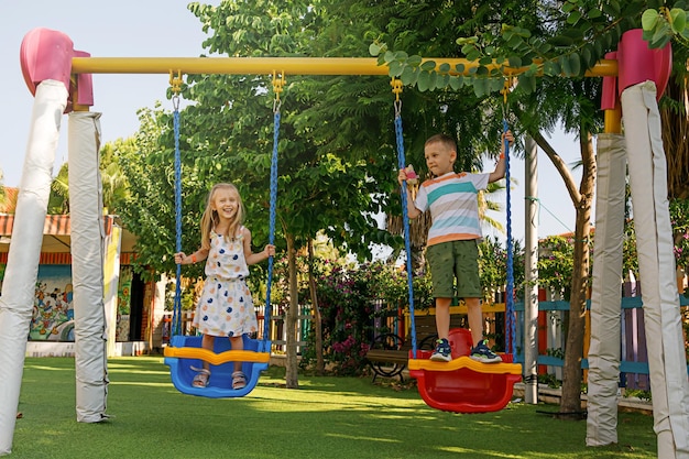 No dia ensolarado de verão, menino e menina balançam no balanço no playground no parque