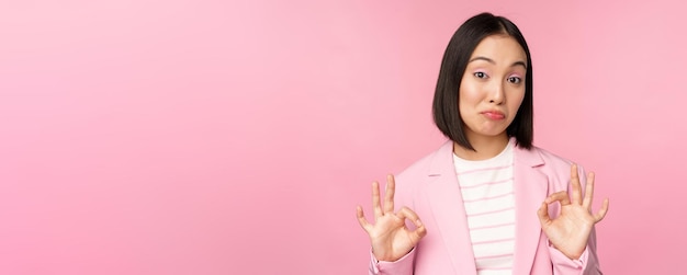 No está mal Una mujer de negocios impresionada, una dama de oficina asiática que muestra un signo de aprobación y asiente con la cabeza, de pie sobre un fondo rosa