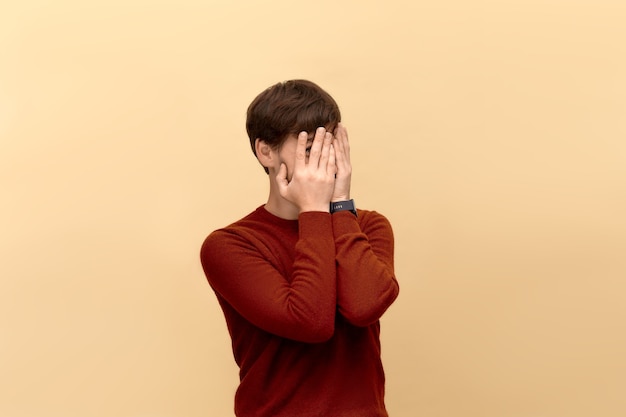 No estoy aquí. Foto de joven tímido con barba vistiendo suéter, rostro cubierto con palmas y asoma a través de sus dedos, posando contra la pared beige.