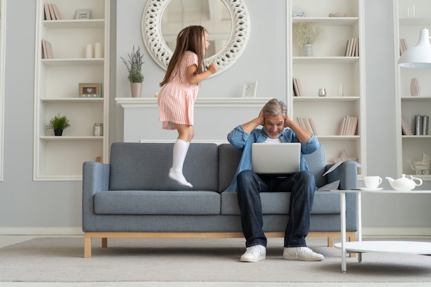 No hay tiempo para los niños. Papá canoso ocupado con la computadora portátil, trabajando en línea en casa, triste hija ofendida aburrida saltando cerca.