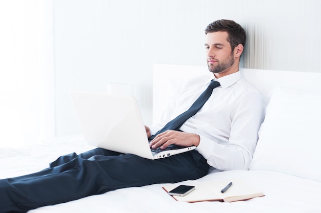 No hay tiempo para relajarse. Hombre joven confidente en camisa y corbata trabajando en la computadora portátil mientras está acostado en la cama en la habitación del hotel