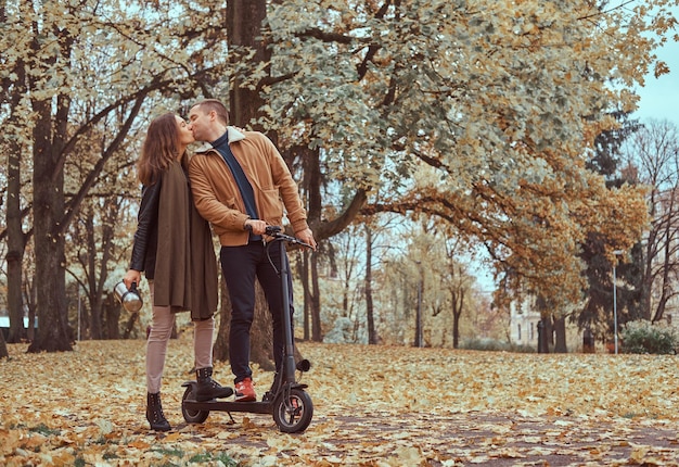 No parque outono cheio de folhas, o homem está beijando uma mulher enquanto segura a scooter.