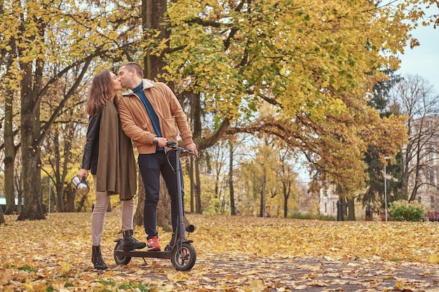 No parque outono cheio de folhas, o homem está beijando uma mulher enquanto segura a scooter.