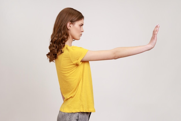 Foto no, prohibido. vista lateral de una joven atractiva y molesta con una camiseta informal amarilla que muestra un gesto de parada, una prohibición o una expresión de advertencia. disparo de estudio interior aislado sobre fondo gris.