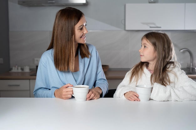 No quadro, uma mãe e uma filha alegres estão sentadas juntas na mesa da cozinha bebendo chá quente pela manhã Menina curiosa pergunta algo à mãe