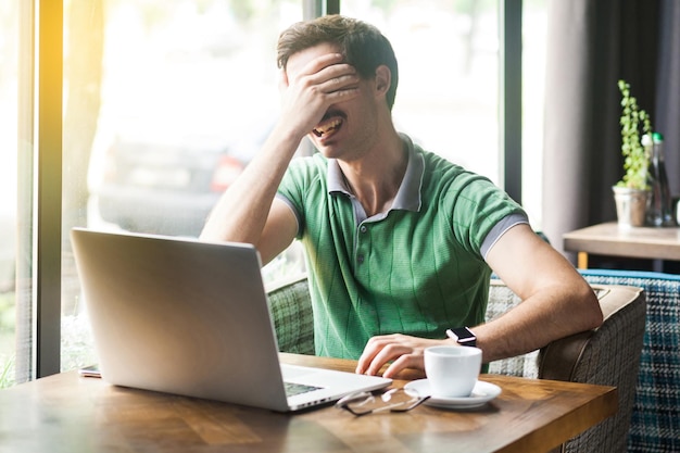 No quiero mirar a este joven hombre de negocios asustado o tímido con una camiseta verde sentado y trabajando en una computadora portátil que cubre sus ojos concepto de negocio e independiente tiro interior cerca de la ventana durante el día