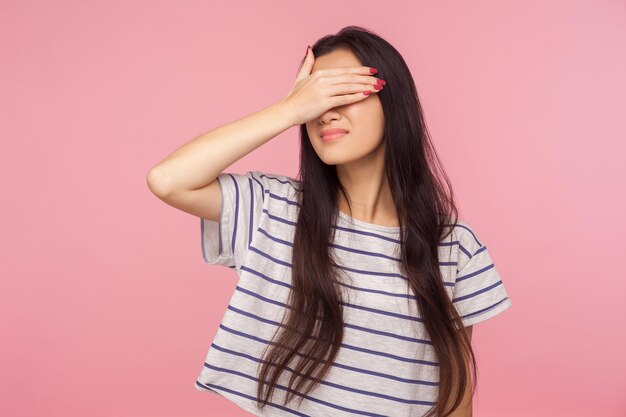 No quiero mirar, Retrato de una chica confundida con el pelo largo en una camiseta a rayas escondiendo los ojos, negándose a mirar vergonzosos eventos malos, ignorando los problemas. tiro de estudio interior aislado sobre fondo de color rosa