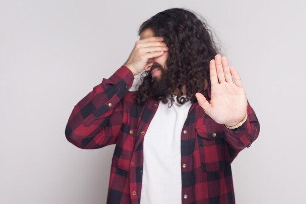 No quiero ver esto. Retrato de hombre guapo con barba y pelo largo y rizado negro en camisa roja a cuadros de pie cubriendo los ojos y gesto de parada. tiro del estudio de interior, aislado en fondo gris.