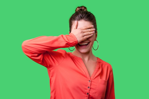 No quiero ver el retrato de una mujer elegante y positiva con peinado de moño, aretes grandes y blusa roja de pie cubriendo los ojos con la mano y una foto de estudio interior sonriente aislada en un fondo verde