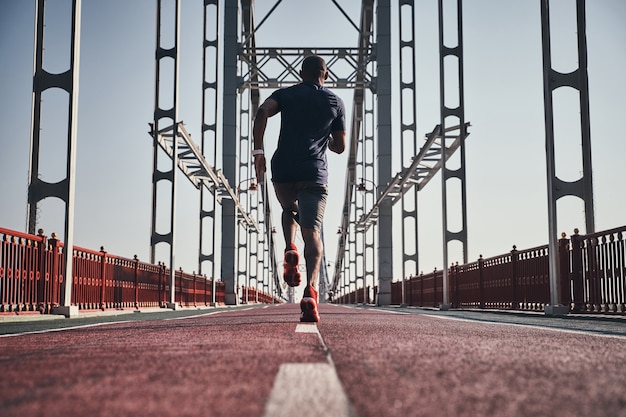 No rendirse nunca. Vista trasera de la longitud completa del joven africano en ropa deportiva haciendo ejercicio mientras trota en el puente al aire libre