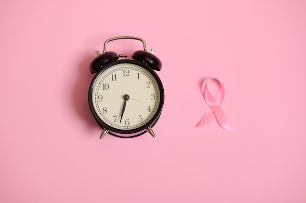 No te pierdas el tiempo, composición plana con reloj despertador y cinta rosa. Concienciación sobre el cáncer de mama, concepto médico aislado sobre fondo rosa con espacio de copia. Concepto de salud de la mujer.