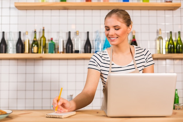 No trabalho. Mulher feliz e positiva encantada em frente ao laptop escrevendo notas enquanto trabalhava no café