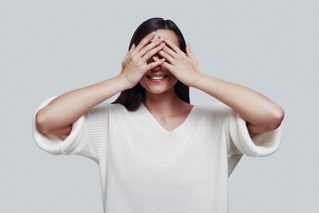 No ver nada. Mujer joven atractiva que cubre los ojos con las manos y sonriendo mientras está de pie contra el fondo gris