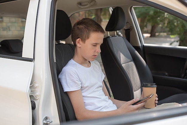 No verão, um adolescente sério de camiseta branca senta no carro com um telefone e um copo de café.