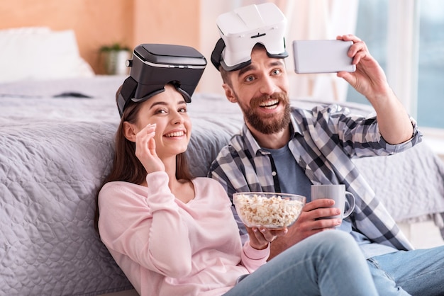 Noch ein Versuch. Fantastisches liebevolles schwules Paar in VR-Gläsern, die glucksen, während sie Selfie nehmen und Popcorn und Kaffee genießen