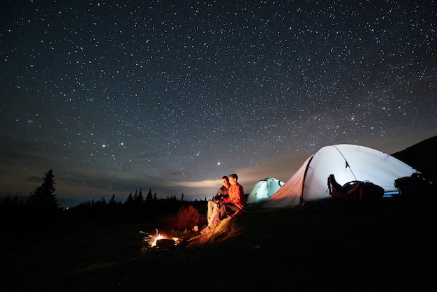 Noche de campamento en las montañas. Turistas hombre y mujer sentados en una fogata cerca de dos carpas iluminadas bajo el cielo estrellado en la noche