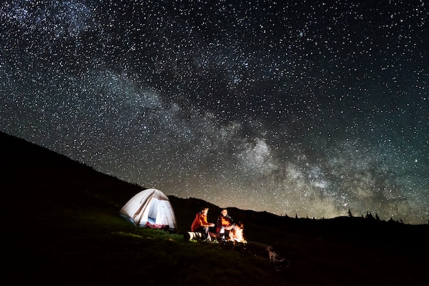 Noche de campamento. Pareja romántica turistas disfrutando en una fogata cerca de una carpa iluminada bajo un hermoso cielo nocturno lleno de estrellas y vía láctea