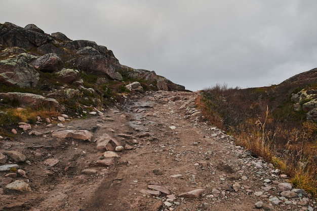 Nördliche Landstraße zwischen Hügeln mit bunten Herbsttundra-Bäumen und Büschen an einem bewölkten Tag. Reise nach Teriberka. Kola-Halbinsel, Murmansk-Region, Russland.