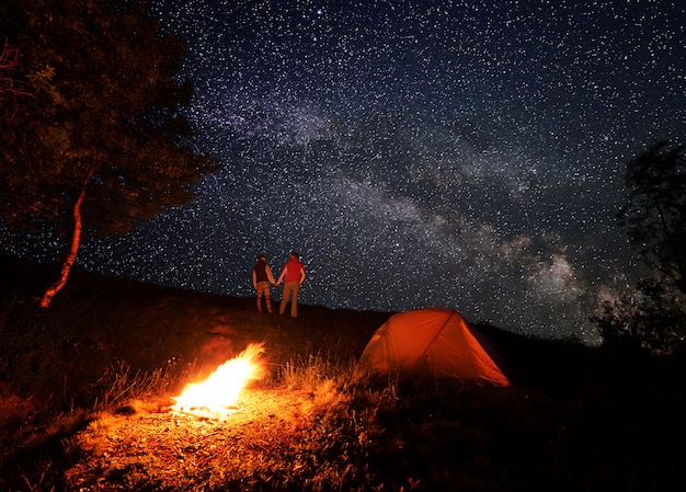 Noite acampando com uma fogueira e barraca sob um céu estrelado e a Via Láctea. Turistas de homem e mulher estão de mãos dadas e se olhando sob o céu romântico nas montanhas