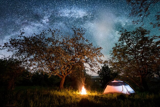 Noite de acampamento perto da cidade. barraca do turista iluminada perto da fogueira sob árvores e lindo céu noturno cheio de estrelas e via láctea