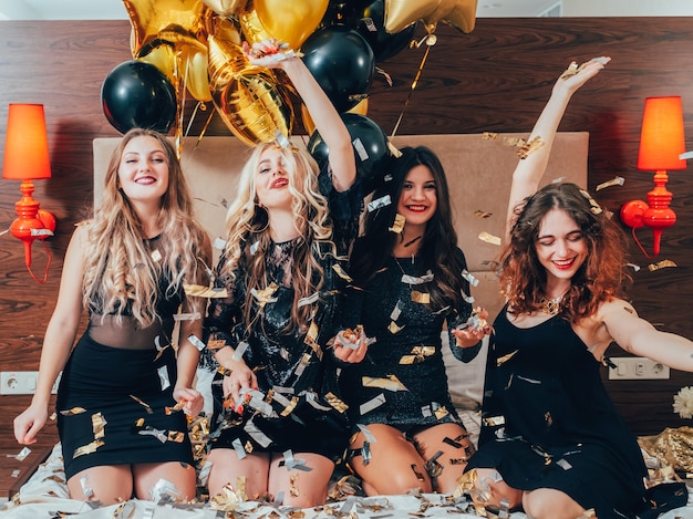 Noite de mulheres fora. Tempo de lazer de festa. Mulheres sorridentes na cama posando. Alegria e liberdade da juventude. Confetes e balões de brilho.