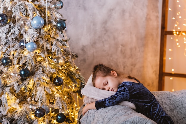 Noite de Natal. Menina cansada adormeceu no sofá enquanto esperava o Papai Noel com presentes no abeto decorado em casa.