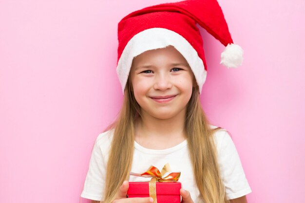 Noite de Natal! Uma criança alegre com um chapéu de Natal em um fundo rosa com uma caixa de presente vermelha.