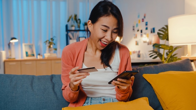 À noite, linda senhora asiática sorridente usando o celular, pedido de um produto de compra online com cartão de crédito no sofá