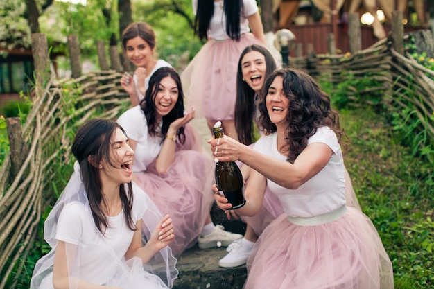 Noiva alegre e suas melhores amigas sentadas em degraus de madeira ao ar livre perto do restaurante. Meninas vestindo camisetas brancas e saias rosa claro. Uma garota abrindo garrafa de champanhe