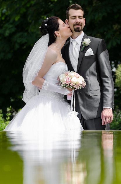 Noiva beijando o noivo enquanto segura flores ao lado da lagoa