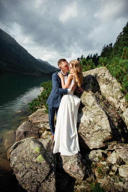 Noiva com lindo vestido branco e noiva com vista para belas montanhas verdes e lago com água azul