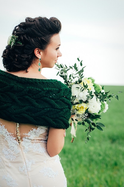 Noiva com um buquê em um vestido de marfim e um xale de tricô em um campo verde