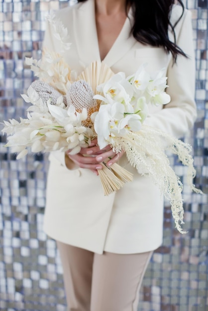 Noiva de terno bege segura buquê boho de casamento com orquídeas banksia e flores secas em um fundo de textura de lantejoulas prateadas