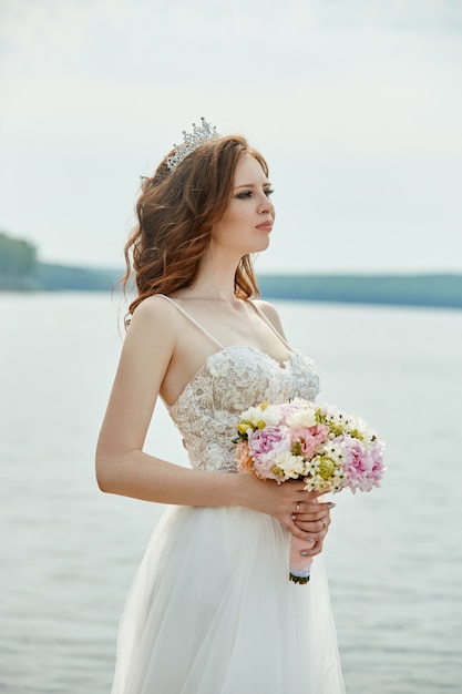 Noiva de vestido branco fica na ponte e olha para o lago. mulher em um vestido de noiva com um buquê de flores