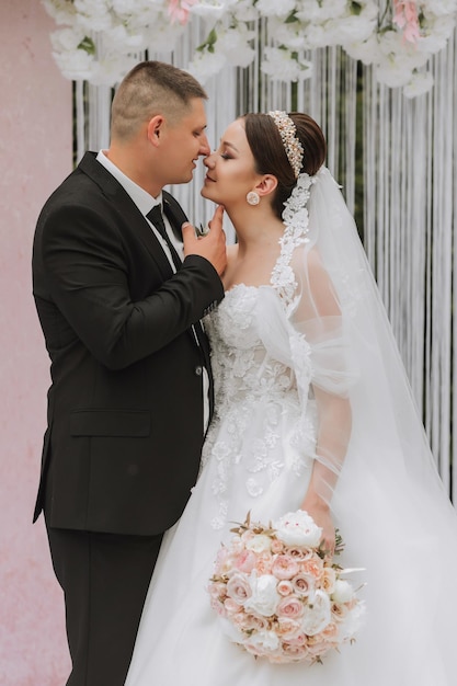 Noiva e noivo atraentes na cerimônia no dia do casamento com um arco feito de flores cor-de-rosa e brancas Belos recém-casados uma jovem mulher em um vestido branco com um longo trem homens em um terno preto