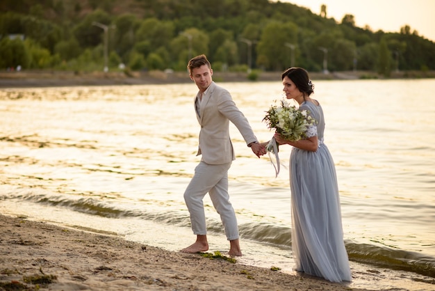 Noiva e noivo com um buquê de casamento caminhando na praia ao pôr do sol