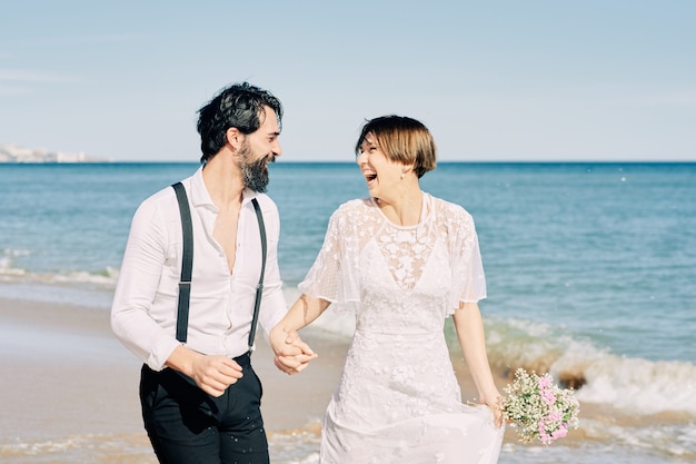 Noiva e noivo correndo ao longo da praia de mãos dadas e sorrindo alegremente, comemorando seu casamento.