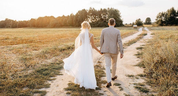 Noiva e noivo no dia do casamento se abraçando e caminhando pela estrada para a floresta ao pôr do sol