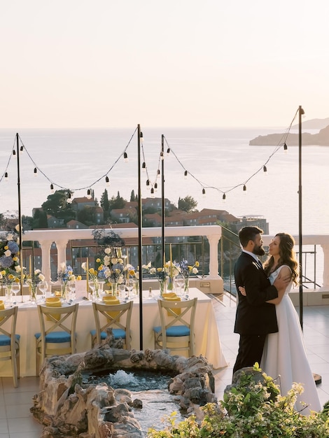 Noiva e noivo se abraçam no terraço perto da mesa posta com vista para a ilha sveti stefan