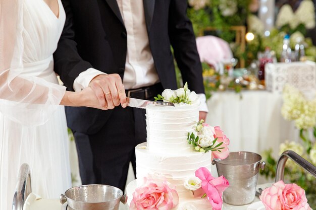 Noiva e noivo segurando uma faca e cortando um elegante bolo de casamento branco com flores