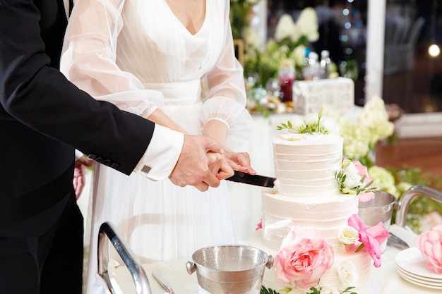 Noiva e noivo segurando uma faca e cortando um elegante bolo de casamento branco com flores