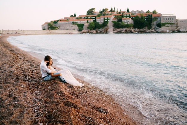 Noiva e noivo sentados em um abraço na praia, tendo como pano de fundo a ilha Sveti Stefan