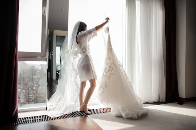 Noiva em roupão e véu segurando o vestido de noiva em um cabide