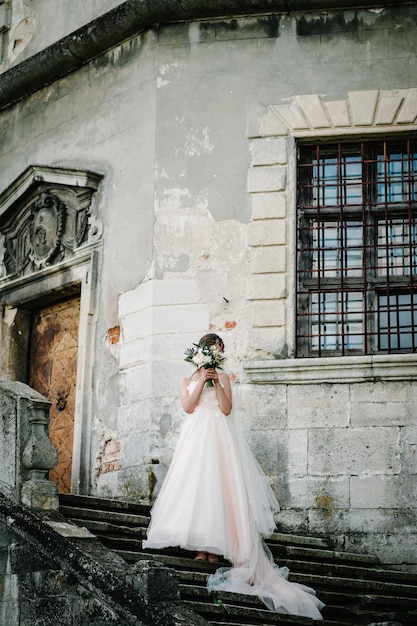 Noiva em vestido de noiva com um buquê de flores fica nas escadas perto da parede arquitetura antiga edifício antigo