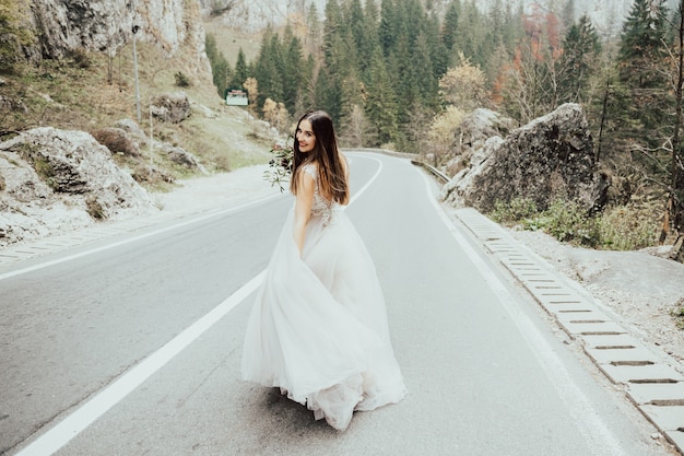 Noiva feliz com um buquê de flores, caminhando ao longo de uma estrada de montanha.