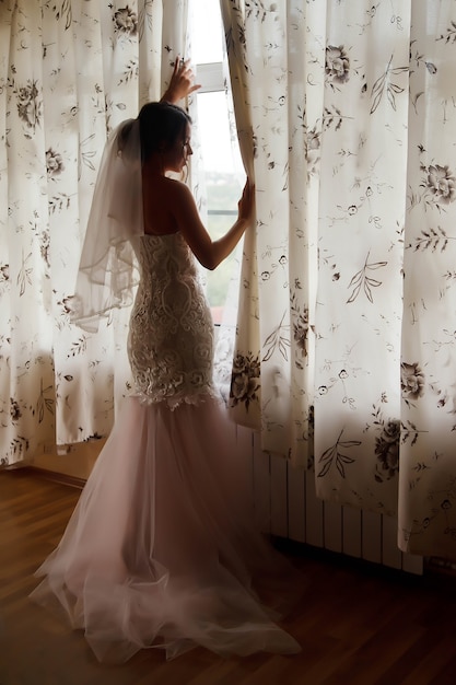 Noiva jovem bonita em casa vestido branco na janela em casa de campo. Manhã da noiva no dia do casamento. Mulher feliz está esperando para conhecer o noivo. Conceito de feliz e luxo casado. Copie o espaço para o site