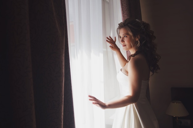 Noiva jovem bonita em um vestido branco na janela em um quarto de hotel. Manhã da noiva no dia do casamento. Mulher feliz está esperando para conhecer seu noivo. Conceito de feliz e luxo casado. Copie o espaço